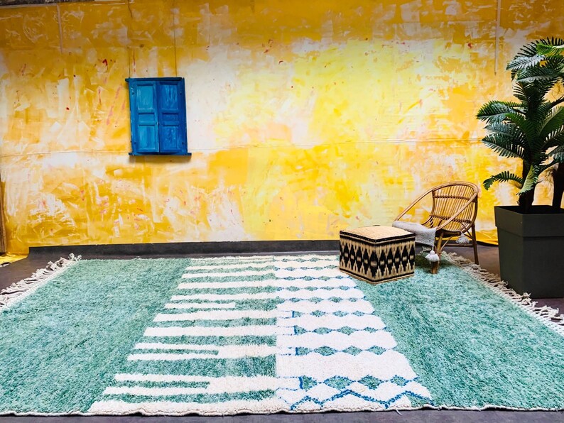 May include: A large, rectangular rug with a teal and white geometric design. The rug features a textured, shaggy pile and a fringe along the edges. The design includes stripes and diamond shapes. The rug is placed on a dark surface against a yellow wall.