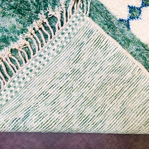 May include: A close-up of a green and white rug with a geometric pattern. The rug has a textured surface and a fringe along one edge. The underside of the rug is visible, showing a striped pattern.