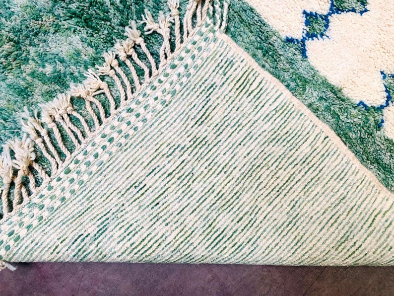 May include: A close-up of a green and white rug with a geometric pattern. The rug has a textured surface and a fringe along one edge. The underside of the rug is visible, showing a striped pattern.