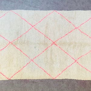 May include: A white rug with a diamond pattern in pink. The rug has a fringe border.