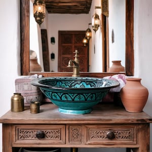 May include: A teal and black patterned ceramic sink with a brass faucet sits atop a wooden vanity with carved drawer fronts. The vanity holds small brass containers and a terracotta vase. A mirror reflects the room's decor.