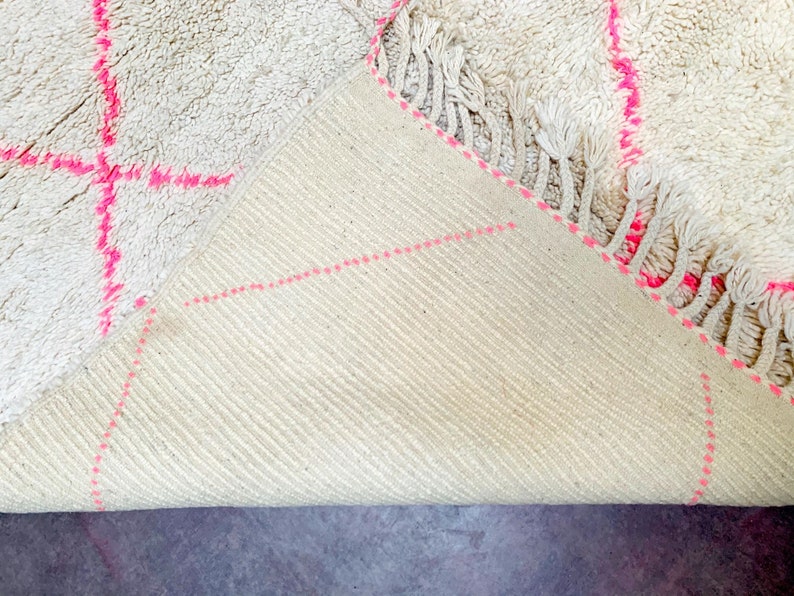 May include: A white rug with pink stitching, folded to show the underside. The rug has a fringe border.