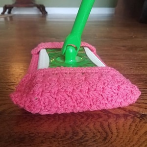 May include: A pink crocheted mop head cover attached to a green mop handle. The cover is made of a textured, hand-stitched yarn. The mop is resting on a wooden floor.