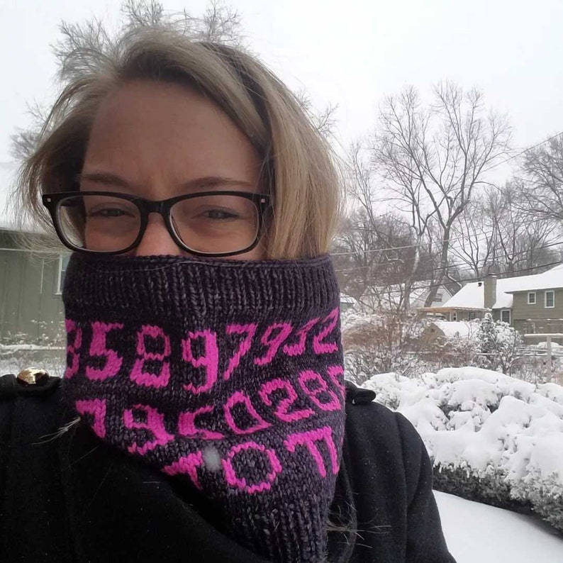 May include: A woman wearing a black knit neck warmer with pink numbers on it. The neck warmer is covering her mouth and nose. She is wearing glasses and has blonde hair. The background is a snowy landscape.