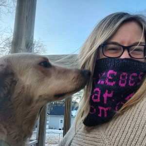 May include: A woman with long blonde hair is wearing a black knitted neck warmer with pink lettering that says "We go hard" and black framed glasses. She is smiling and looking at a golden brown dog.