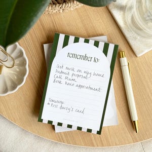May include: A green and white striped notepad with a rounded white section at the top. The text "remember to:" is written in black at the top of the white section. The notepad has a handwritten to-do list that includes "Get milk on way home", "Submit proposal", "Call Mum", and "Book hair appointment". The text "Tomorrow:" is written in black below the to-do list, followed by a handwritten note that says "*Post Daisy's card".