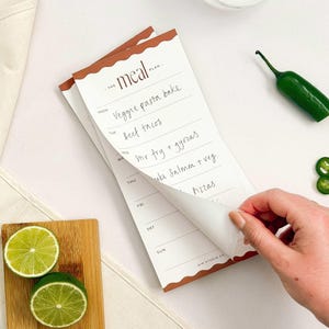 May include: A white paper meal planner notepad with a brown scalloped border at the top and bottom. The header reads THE MEAL PLAN. The days of the week are listed vertically with handwritten entries including veggie pasta bake and beef tacos.