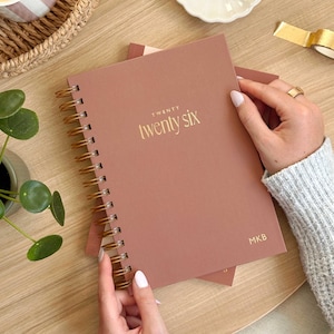 May include: A dusty rose spiral-bound planner with gold lettering that reads "TWENTY twenty six". The planner is held by a person. The initials "MKB" are printed in gold on the bottom right corner.