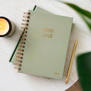 Op de afbeelding: Een saliegroene spiraalplanner met gouden draadbinding. De kaft bevat de goudfolie tekst twenty seven en de initialen MKB onderaan. Een gouden pen ligt naast de planner, die bij een brandende kaars in een zwarte pot is geplaatst.