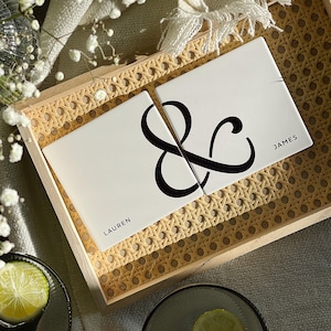 Puede incluir: Dos posavasos de cerámica blanca con un símbolo de ampersand negro y los nombres Lauren y James. Los posavasos están en una bandeja tejida con un marco de madera claro.