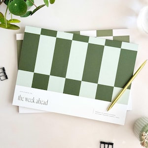 Può includere: Un blocco per pianificazione settimanale rettangolare con un motivo a scacchi verde e verde chiaro. La sezione bianca inferiore mostra il testo "AIM STUDIO CO", "the week ahead" e "MONDAY TO SUNDAY PLANNER WITH HABIT TRACKER & WEEKLY GOAL". Una penna dorata poggia sulla carta.