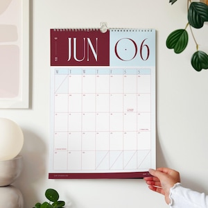 a woman's hand is turning the page on a large wall calendar showing the month of June 2027 in a deep red and sky blue colour palette.