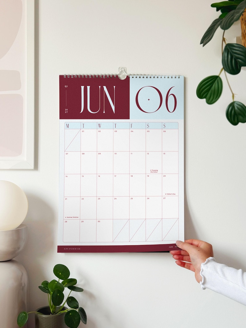 a woman's hand is turning the page on a large wall calendar showing the month of June 2027 in a deep red and sky blue colour palette.