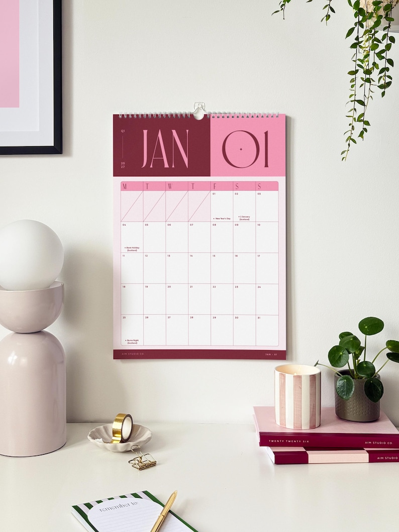 Styled home office desk showing a bold 2027 wall calendar hanging on the wall, surrounded by minimal wall art, a hanging plant and stacked notebooks on the desk below.