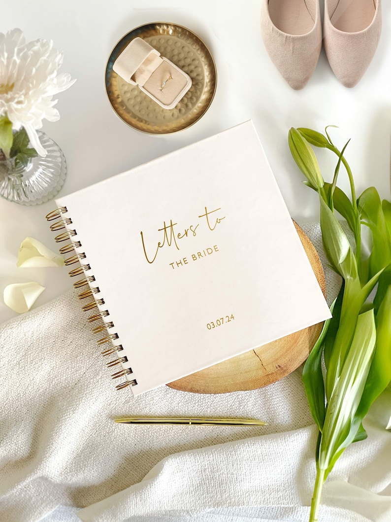 May include: A white spiral bound journal with the gold text "Letters to the Bride" and the date "03.07.24". The journal is on a wooden coaster and a gold pen is next to it.