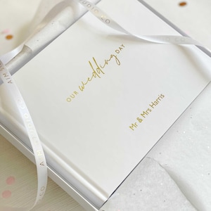 a first anniversary gift for wife wrapped elegantly in white tissue paper with ribbon and gift box revealing a personalised memory book with "our wedding day" in gold foil on the cover.