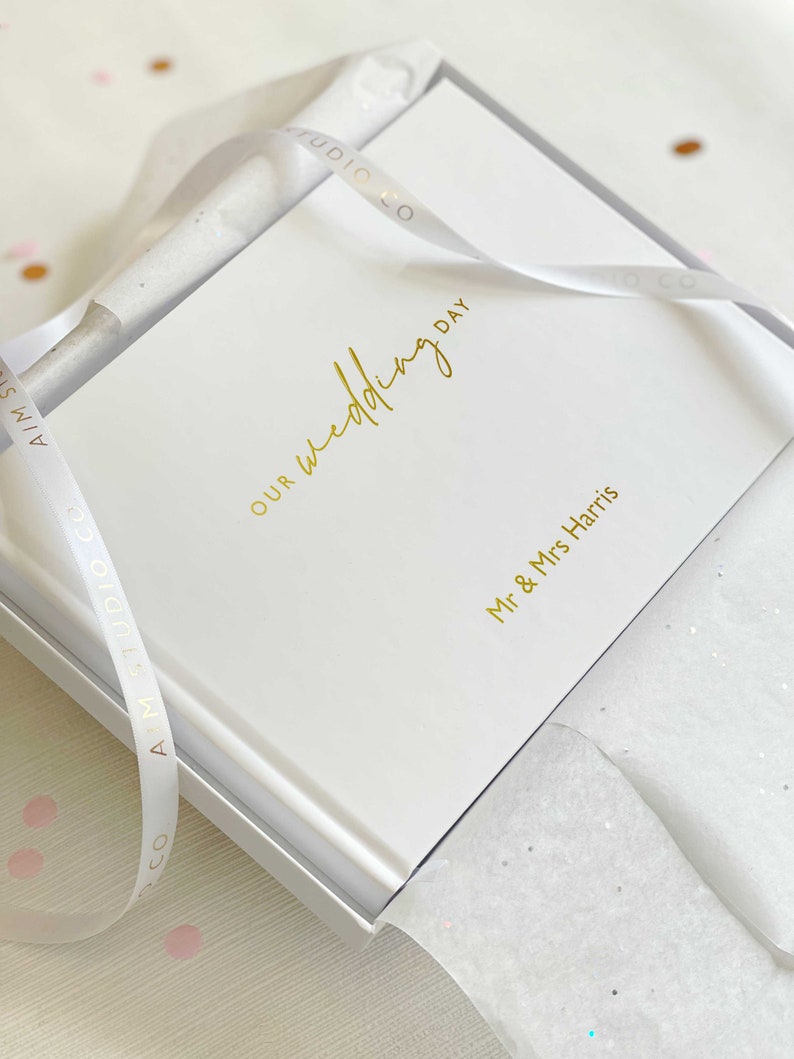 a first anniversary gift for wife wrapped elegantly in white tissue paper with ribbon and gift box revealing a personalised memory book with "our wedding day" in gold foil on the cover.