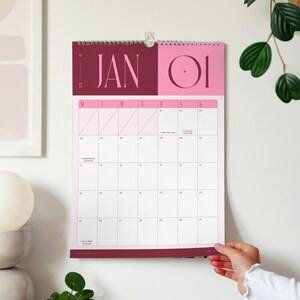 A spiral-bound A3 monthly calendar hanging on a wall showing the month of January 2027 with colourful pink and burgundy heading and spacious minimal planner grid.