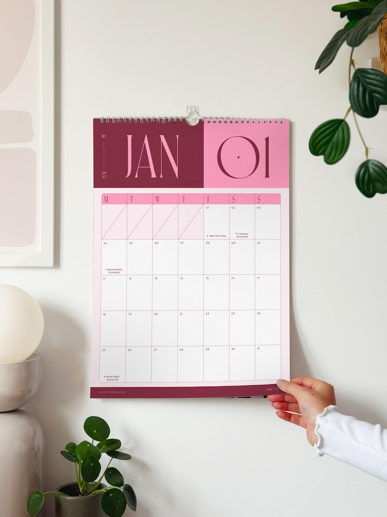 A spiral-bound A3 monthly calendar hanging on a wall showing the month of January 2027 with colourful pink and burgundy heading and spacious minimal planner grid.