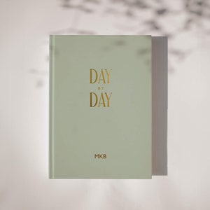 Peut inclure: Un journal vert clair avec le texte "DAY BY DAY" en lettres dorées et "MKB" en lettres dorées en bas.