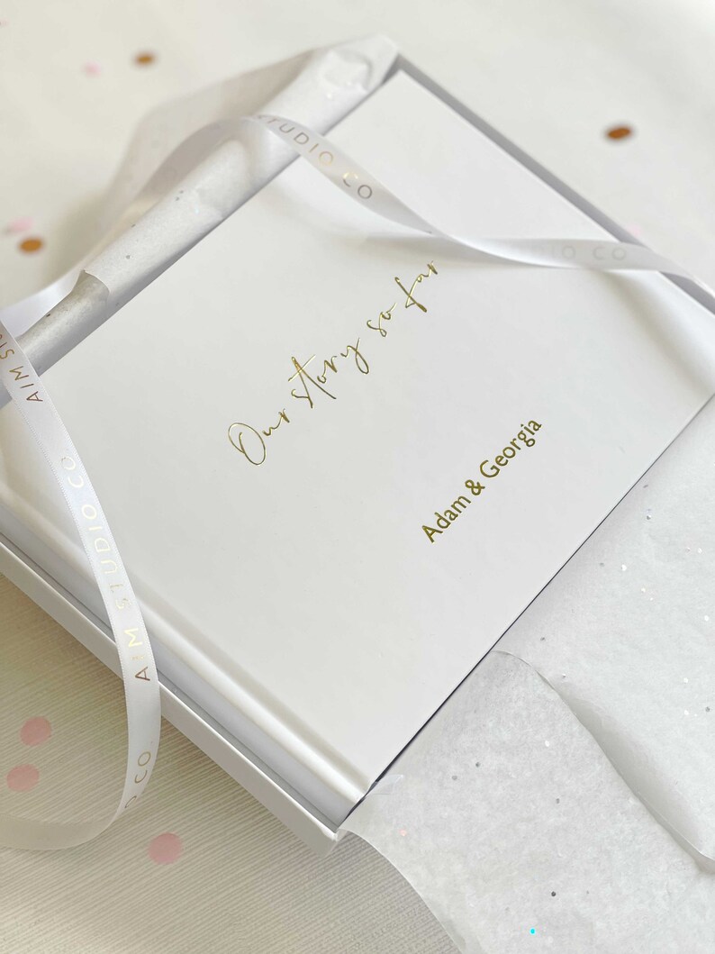 a white scrapbook in gift box with personalised names of couples and our story so far in gold lettering on the cover.