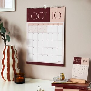 A hanging monthly wall planner showing October 2027 in brown colour way above an office desk. The desk beneath has a matching desk calendar, styled with stacked notebooks and diaries, surrounded by a vase and a candle for a cozy atmosphere.