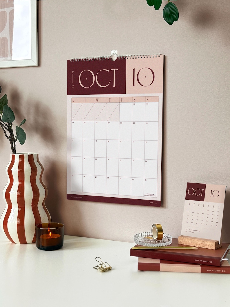A hanging monthly wall planner showing October 2027 in brown colour way above an office desk. The desk beneath has a matching desk calendar, styled with stacked notebooks and diaries, surrounded by a vase and a candle for a cozy atmosphere.