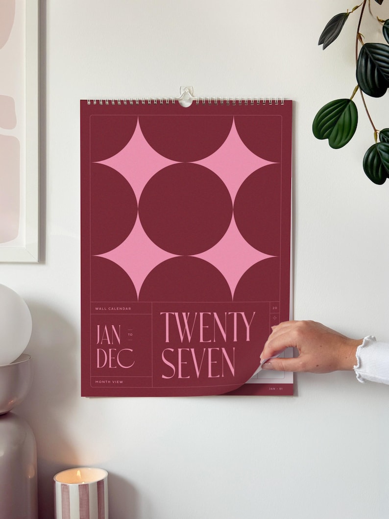 Large wall calendar with a retro burgundy cover, pink stars and '2027' text. Styled on a home office wall, hanging above a desk.