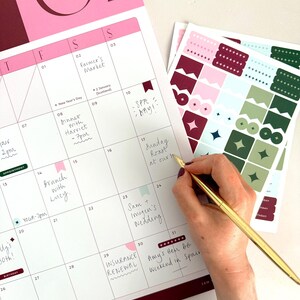 woman writing plans onto january 2027 monthly calendar page with a slim gold pen. The page is decorated with matching planner stickers in coordinating colours.