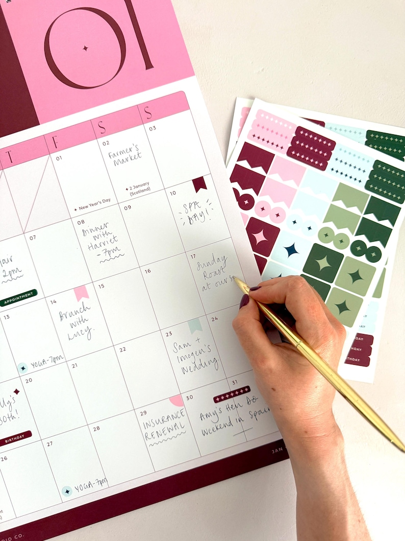 woman writing plans onto january 2027 monthly calendar page with a slim gold pen. The page is decorated with matching planner stickers in coordinating colours.
