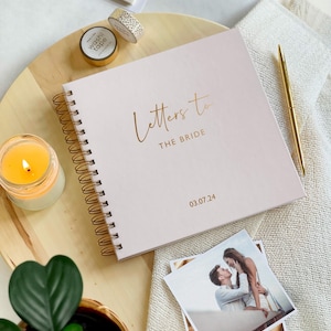 May include: A pink spiral bound journal with the text "Letters to the Bride" and the date "03.07.24" in gold lettering. The journal is on a wooden surface with a candle, a plant, and a photo of a couple.