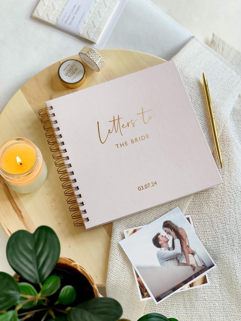 May include: A pink spiral bound journal with the text "Letters to the Bride" and the date "03.07.24" in gold lettering. The journal is on a wooden surface with a candle, a plant, and a photo of a couple.