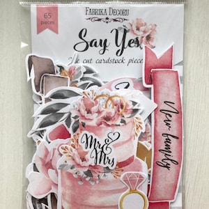 May include: A set of 65 die-cut cardstock pieces with a wedding theme. The pieces feature a pink and white wedding cake with the words "Mr. & Mrs." on it, a ring, flowers, and the words "Say Yes" and "New Family".
