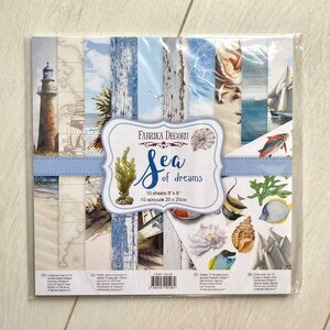 Puede incluir: Un paquete de papel decorativo "Sea of Dreams" de Fabrika Decoru. El paquete contiene 10 hojas de 20 cm x 20 cm cada una. El papel presenta diseños de temática náutica, incluyendo un faro, veleros y conchas marinas.