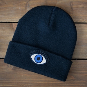 May include: A navy blue beanie hat featuring an embroidered eye design in white and blue on the folded brim. The hat is made of a soft, knitted material. The hat is displayed on a wooden surface.