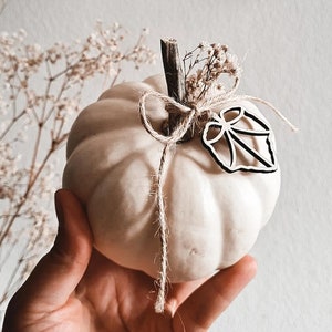Könnte beinhalten: Ein weißer Keramik-Kürbis mit einem zarten Blumenmuster und einem Holz-Blattakzent. Der Kürbis ist mit Schnur gebunden und hat eine rustikale, ländliche Ästhetik.