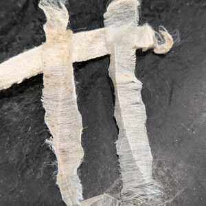 May include: Close-up of a white, frayed fabric strip with a rough texture. The fabric is torn and has a few brown spots. The fabric is on a black background.
