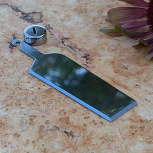 Op de afbeelding: Een zilveren flessenopener met een haakvormig handvat en een scherpe mes. De opener ligt op een houten oppervlak.