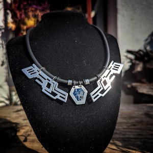 Może przedstawiać: Srebrny naszyjnik z geometrycznymi kształtami i czarnym gumowym sznurkiem. Naszyjnik posiada centralną sześciokątną zawieszkę z niebiesko-srebrnym wzorem. Naszyjnik jest prezentowany na czarnym manekinie.