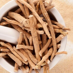 May include: An overhead shot shows a white mortar and pestle filled with light brown ashwagandha roots. A white pestle with a floral design rests inside the mortar. Light brown powder surrounds the mortar on a white surface.