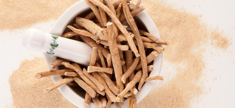 May include: An overhead shot shows a white mortar and pestle filled with light brown ashwagandha roots. A white pestle with a floral design rests inside the mortar. Light brown powder surrounds the mortar on a white surface.