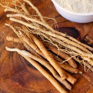 May include: Close-up of ashwagandha roots and powder in a white bowl on a wooden surface. The roots are light brown and beige, with a fibrous texture. A green leaf is visible in the background.