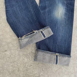 May include: Close-up of blue denim jeans with cuffed hems. The cuffs reveal a lighter gray fabric with a white stripe detail. The jeans show signs of wear and fading, with a distressed edge on one cuff.