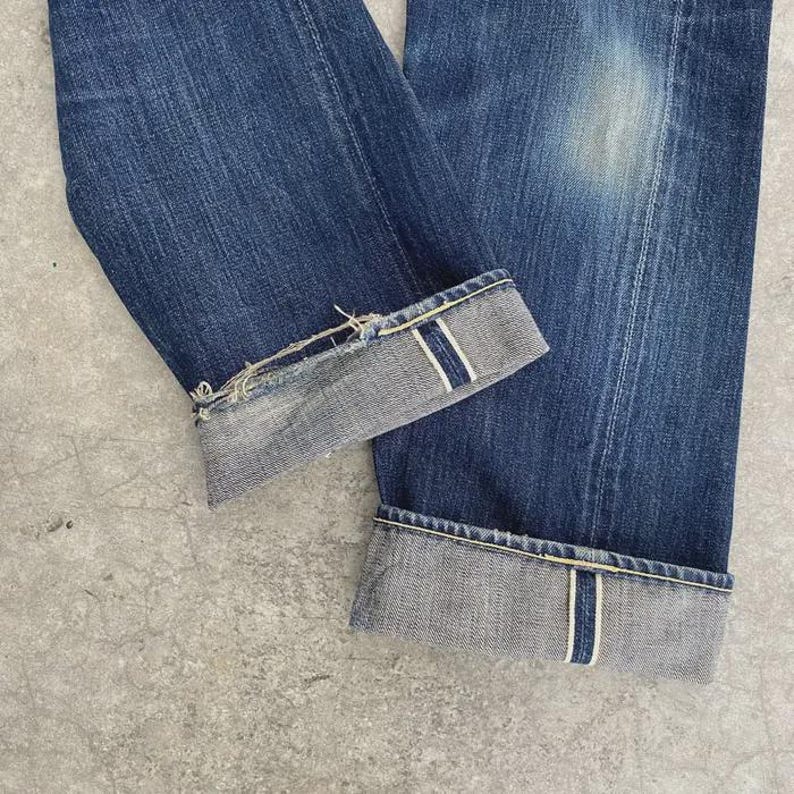 May include: Close-up of blue denim jeans with cuffed hems. The cuffs reveal a lighter gray fabric with a white stripe detail. The jeans show signs of wear and fading, with a distressed edge on one cuff.