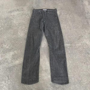 May include: A pair of grey denim jeans with a straight leg design. The jeans have a classic five-pocket style, a button and zip closure, and a textured appearance. The trousers are displayed on a grey concrete surface.