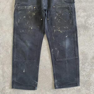 Puede incluir: Pantalones de trabajo gris oscuro con salpicaduras de pintura. Los pantalones tienen refuerzos en las rodillas y m&uacute;ltiples bolsillos. La tela parece desgastada y descolorida. El estilo es utilitario y pr&aacute;ctico.