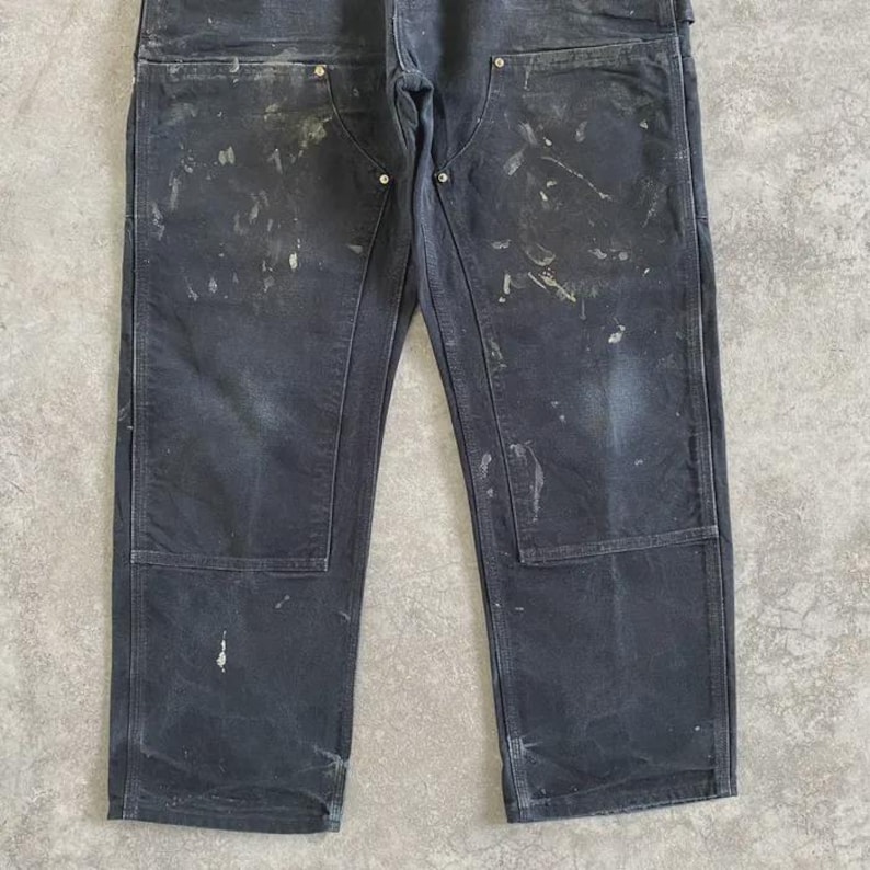 Puede incluir: Pantalones de trabajo gris oscuro con salpicaduras de pintura. Los pantalones tienen refuerzos en las rodillas y m&uacute;ltiples bolsillos. La tela parece desgastada y descolorida. El estilo es utilitario y pr&aacute;ctico.