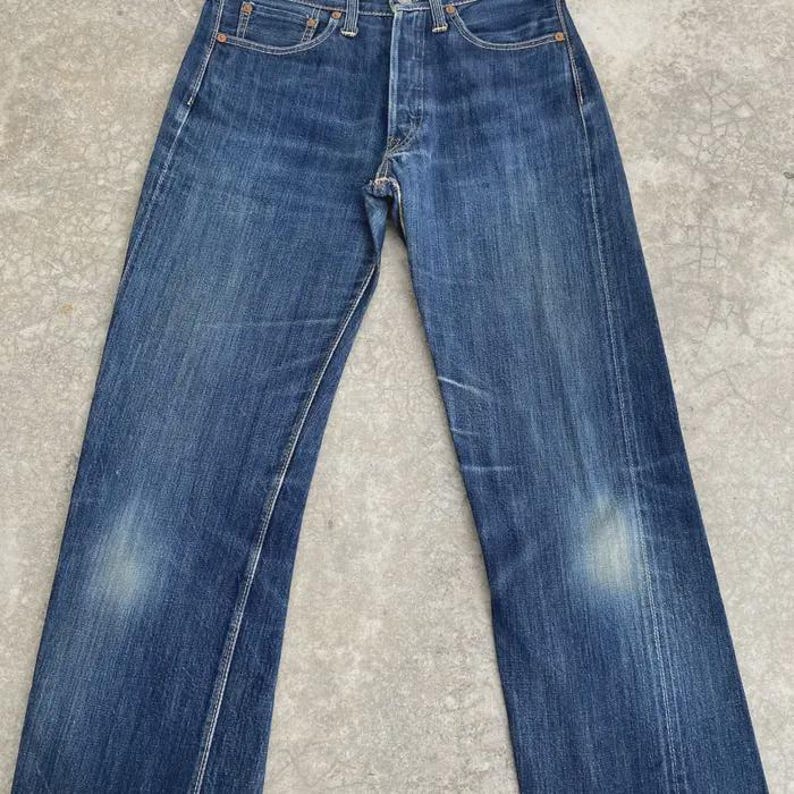 May include: Faded blue denim jeans with a button fly. The jeans have a classic five-pocket design and show signs of wear and fading, with lighter patches on the thighs. The denim has a medium weight and a straight leg cut.