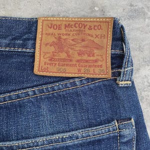 May include: Close-up of blue denim jeans with a brown leather patch. The patch reads "JOE McCOY & CO. ILLINOIS REAL WORK CLOTHING XH" with a graphic of a cowboy on a horse. The jeans are size W28 L36.