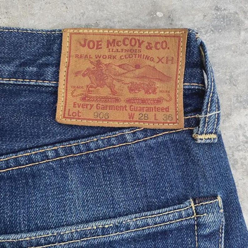 May include: Close-up of blue denim jeans with a brown leather patch. The patch reads "JOE McCOY & CO. ILLINOIS REAL WORK CLOTHING XH" with a graphic of a cowboy on a horse. The jeans are size W28 L36.